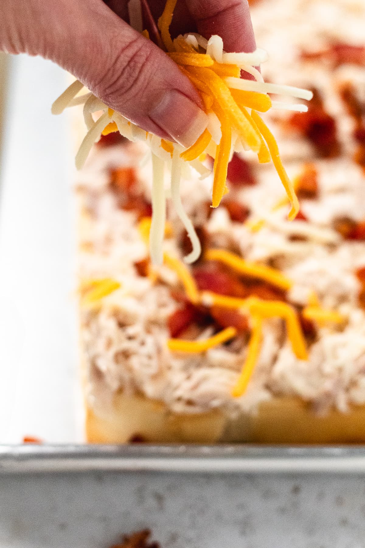 Hand sprinkling shredded cheese over chicken and bacon on Hawaiian roll bottoms.