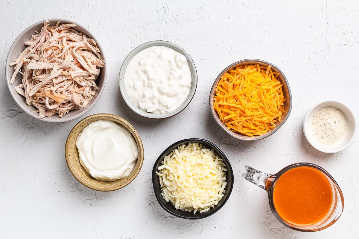 Separate bowls filled with shredded chicken, cheeses, cottage cheese, Greek yogurt, and hot sauce.