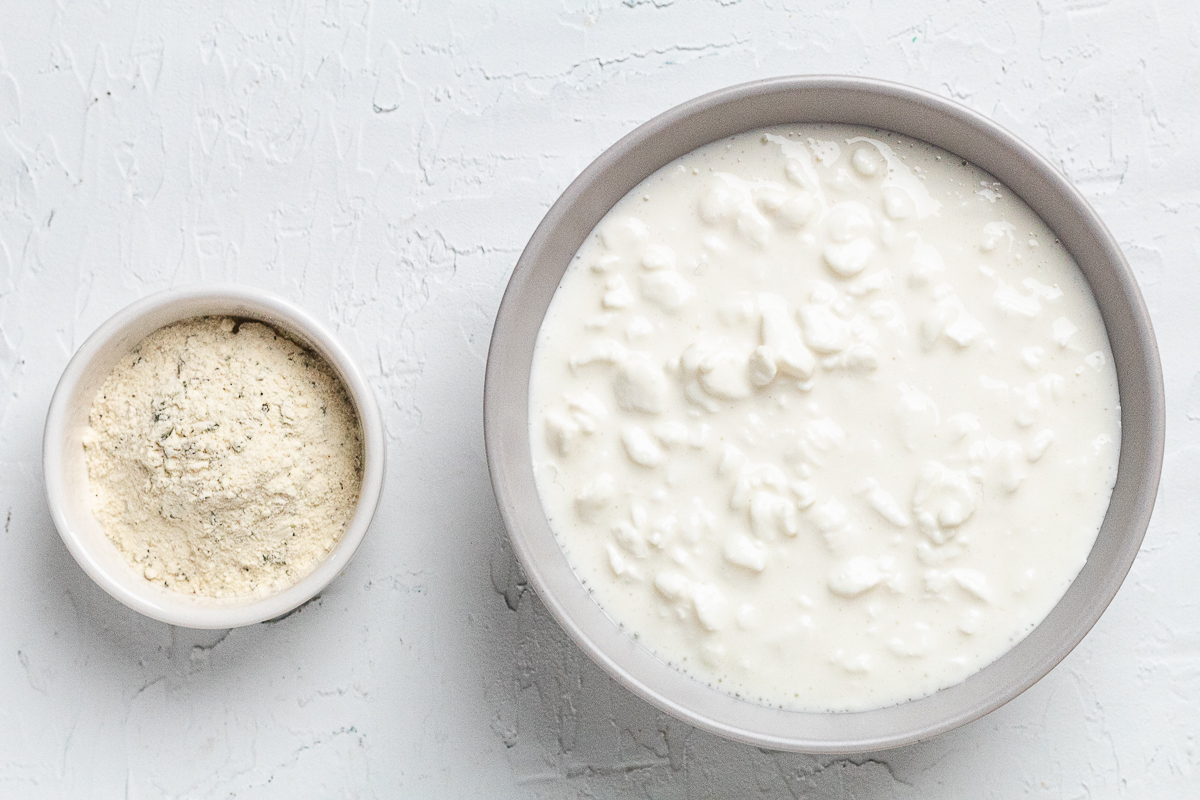 Small bowls of cottage cheese and ranch seasoning on a white surface, ready to make a high protein dip.
