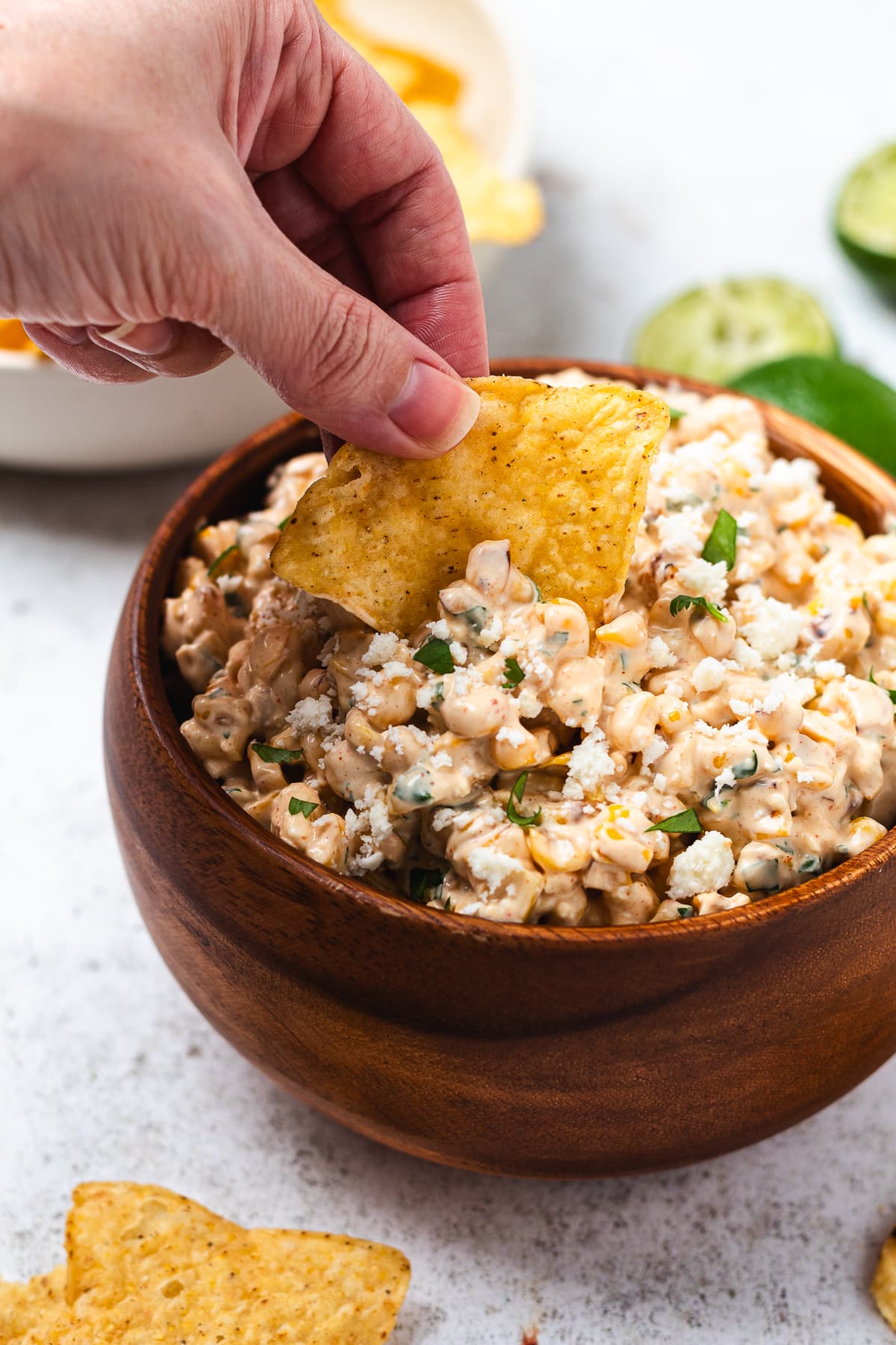 Hand dipping a tortilla chip into creamy corn dip with chips and lime slices nearby.