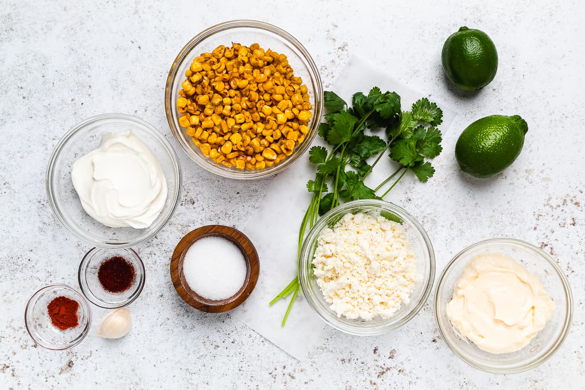 Roasted corn, sour cream, mayo, cotija, spices, garlic, cilantro, and limes arranged on a light surface.