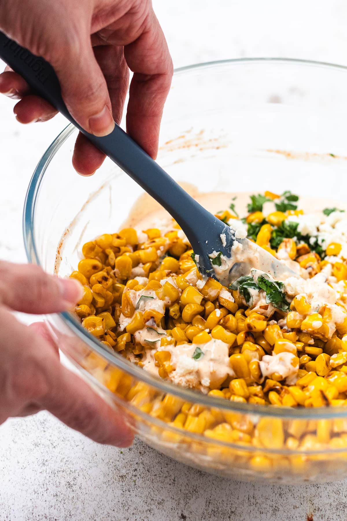 Hand stirring corn, herbs, and creamy sauce in a glass bowl with a blue spatula.