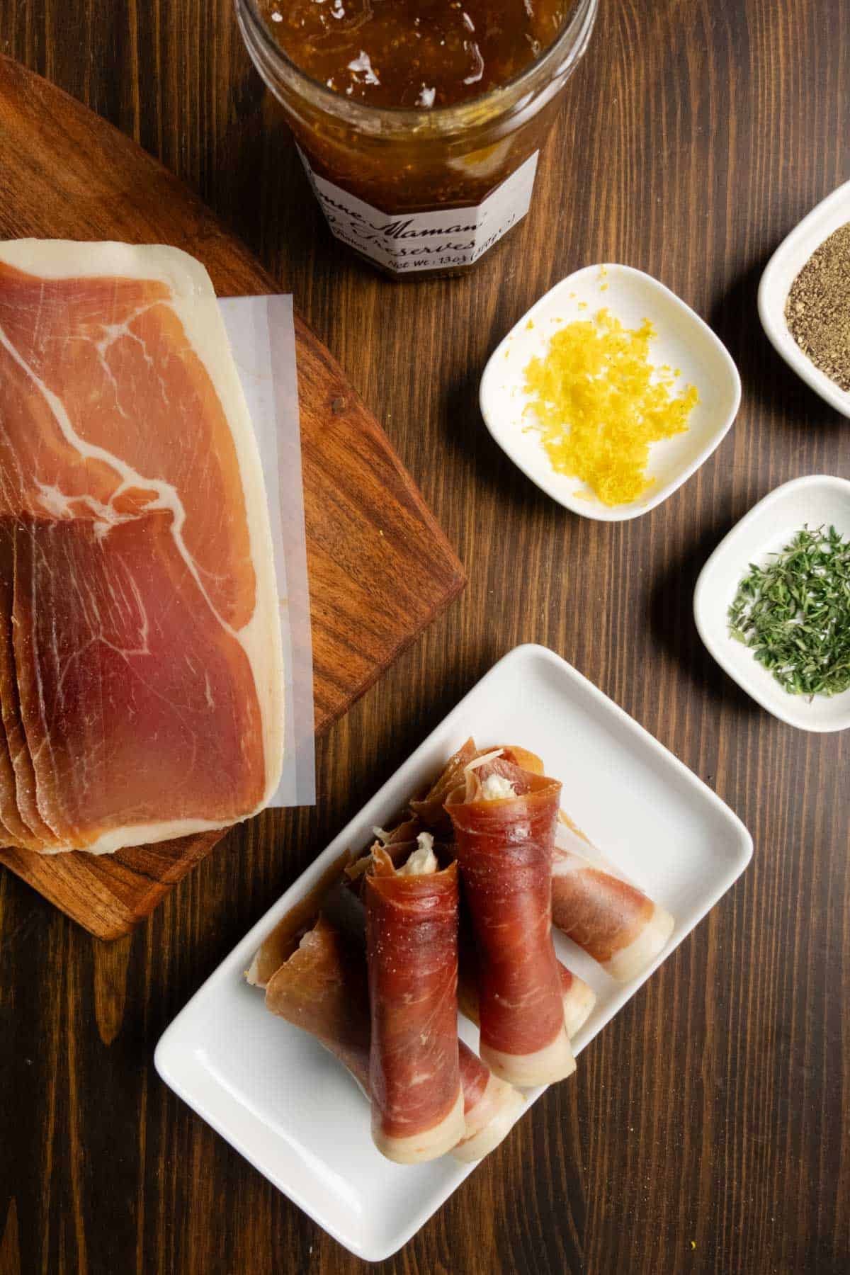 Prosciutto slices and rolled appetizers with fig jam, lemon zest, thyme, and pepper on a wooden board.