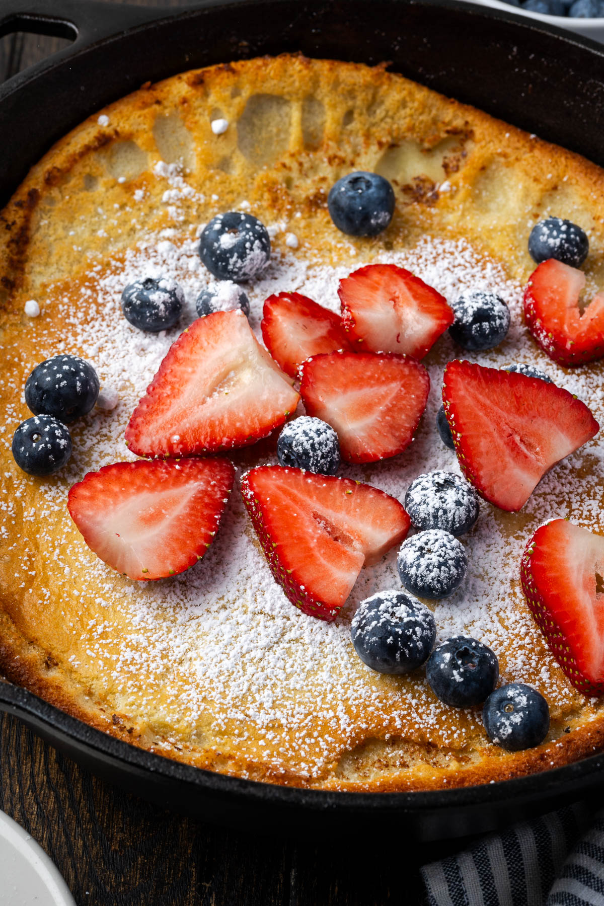 Dutch baby pancake topped with powdered sugar, strawberries, and blueberries in a skillet.