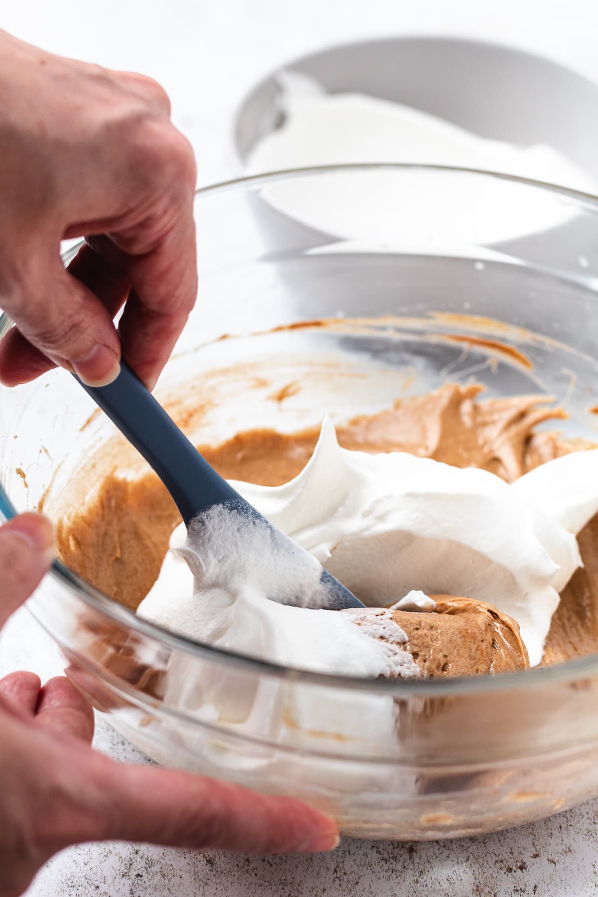 Hands folding whipped white mixture into a light brown mixture with a blue spatula in a glass bowl.
