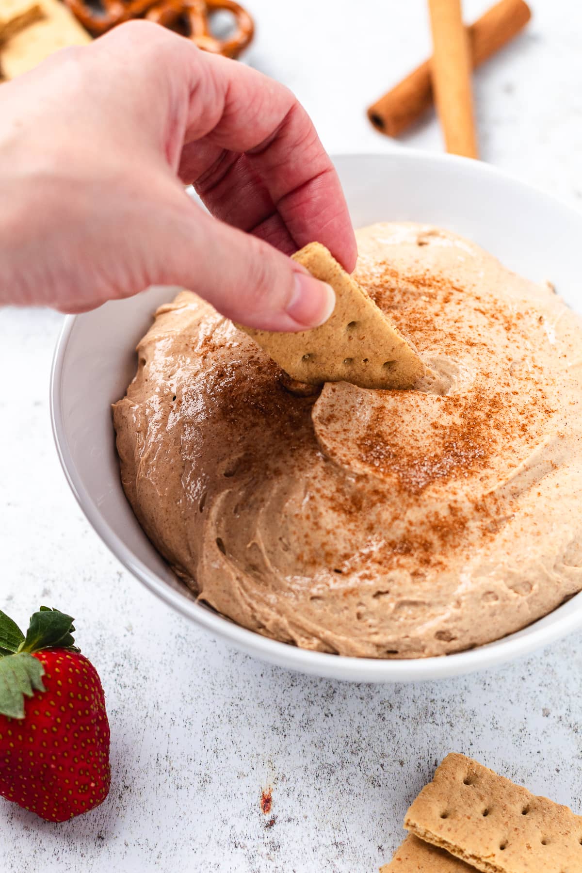 Hand dipping a graham cracker into creamy cinnamon dip with strawberries, pretzels, and crackers nearby.