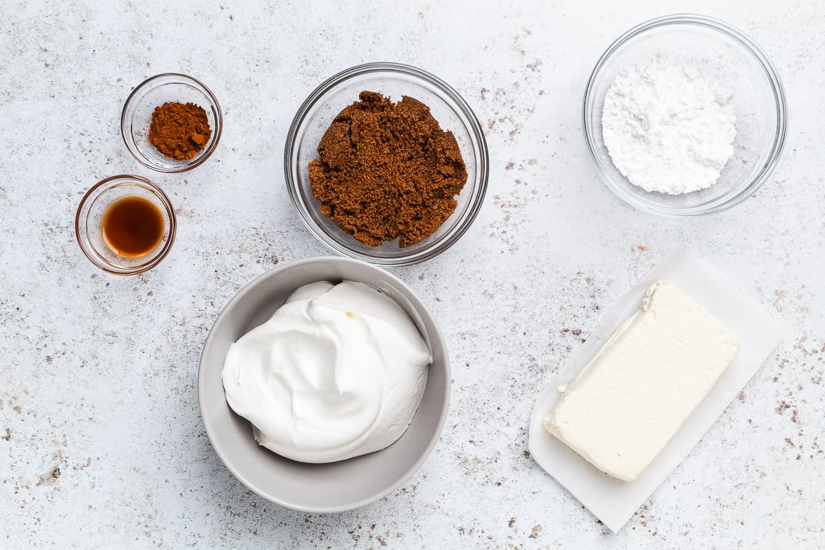 Cinnamon powder, vanilla, brown sugar, powdered sugar, whipped topping, and cream cheese on a light surface.