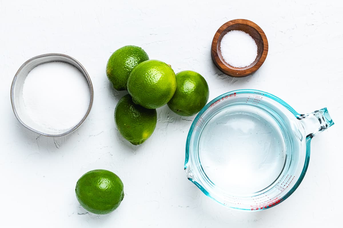 Ingredients for homemade limeade: fresh limes, sugar, salt, and water arranged on a white surface.