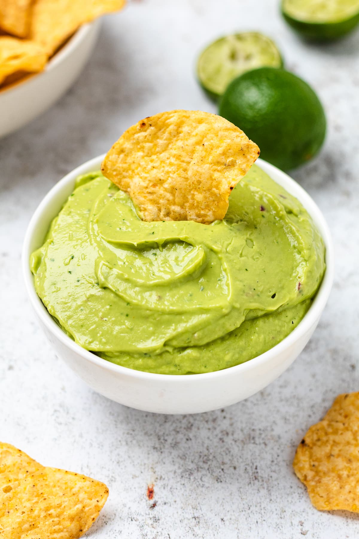 Bowl of smooth green guacamole with a chip standing in it and chips and limes around it.