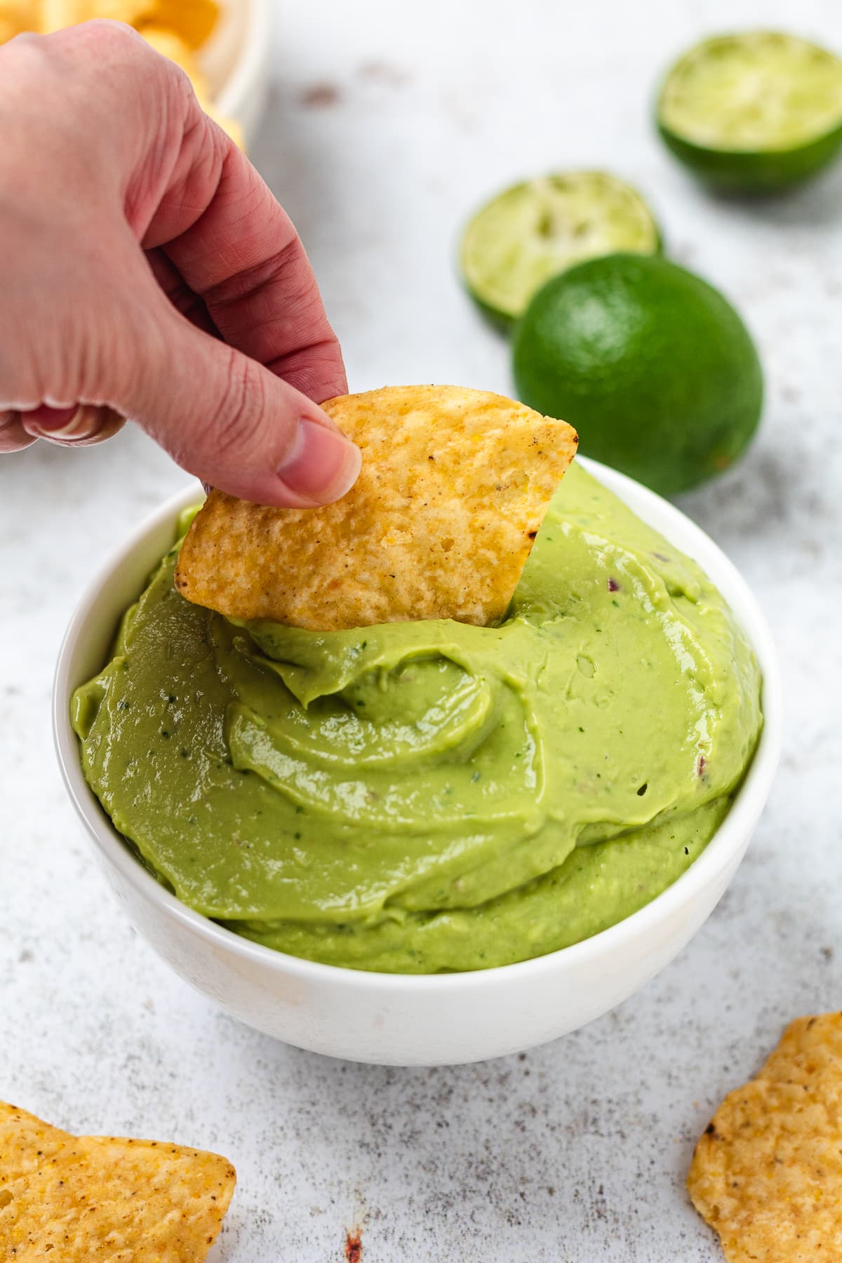 Hand dipping a tortilla chip into creamy green dip with whole and halved limes in the background.