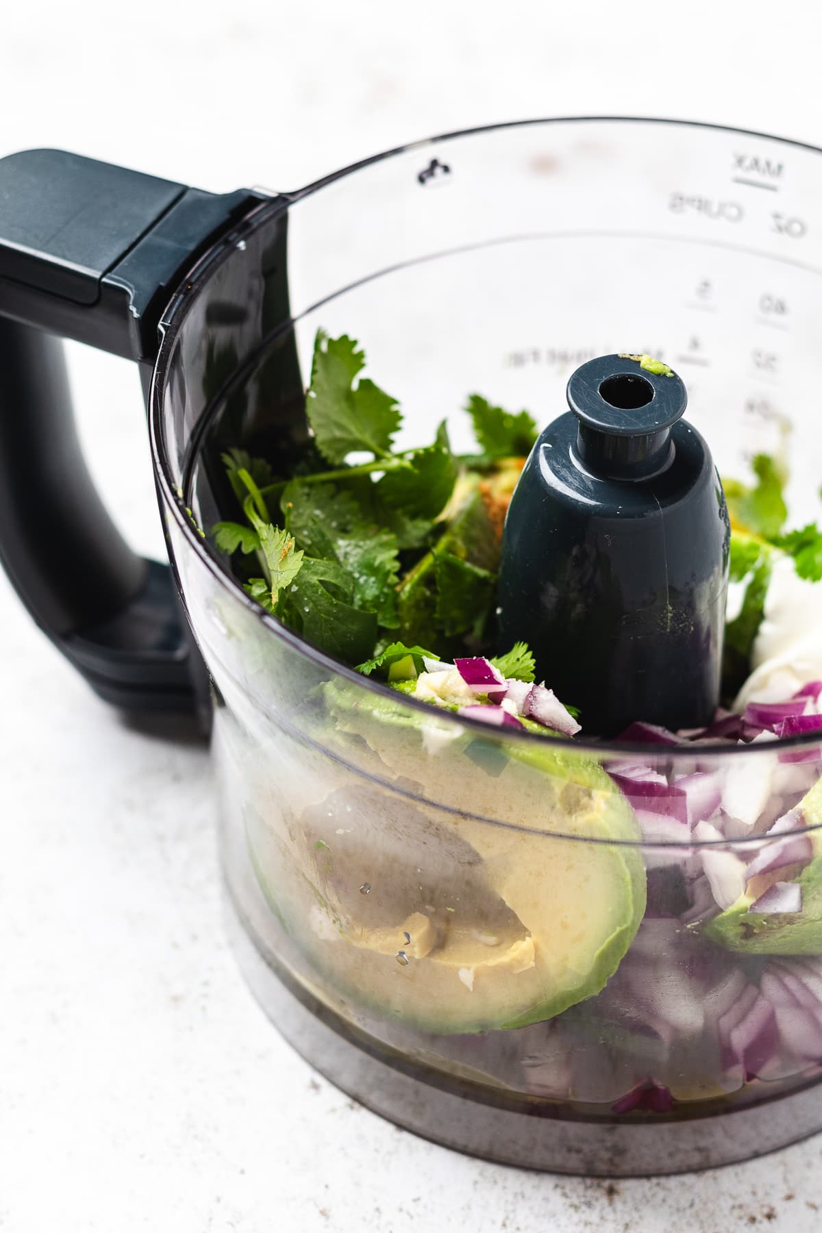 Food processor bowl with avocado, red onion, cilantro, and a dollop of white cream before blending.