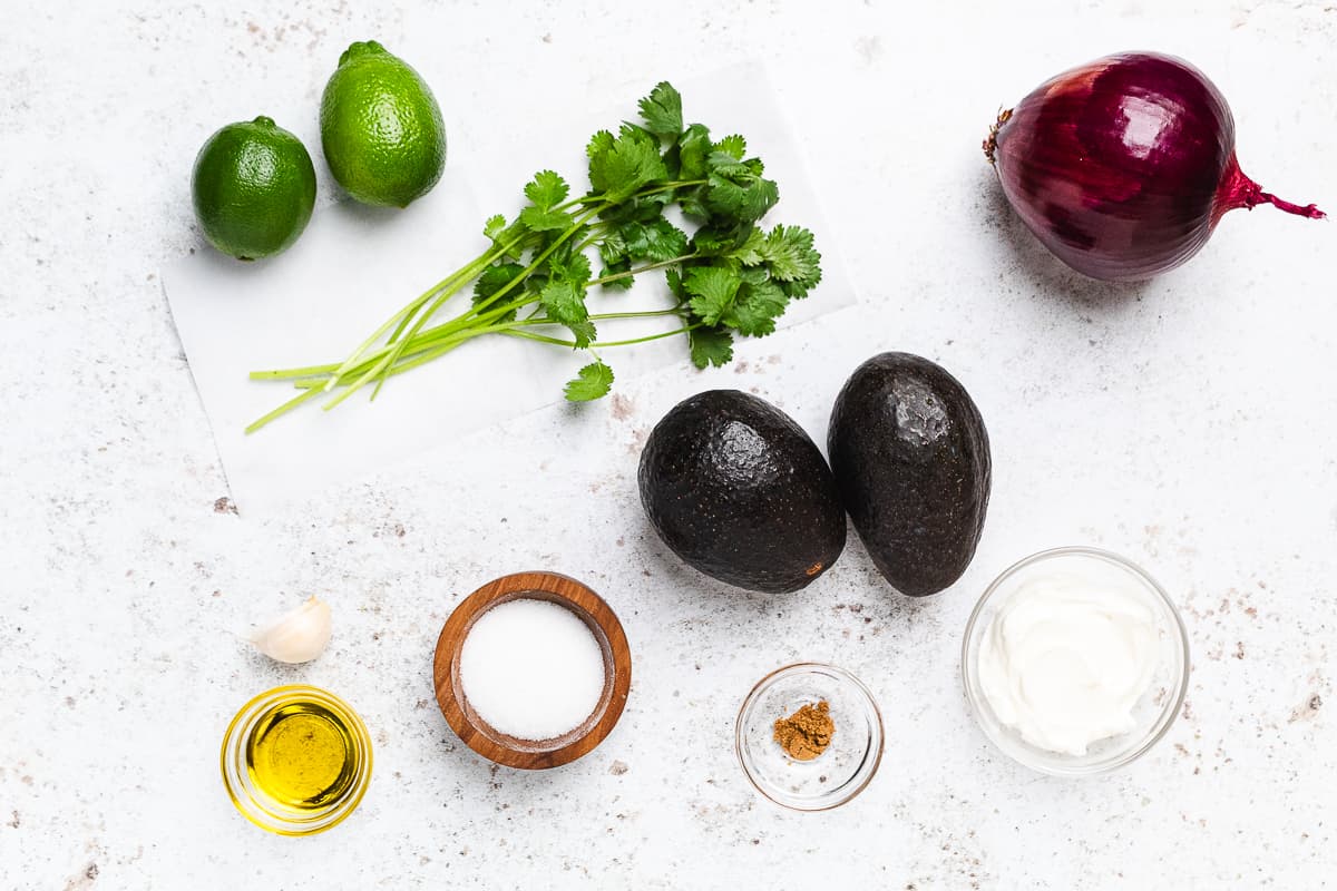 Limes, cilantro, red onion, avocados, garlic, salt, olive oil, cumin, and sour cream arranged on a light surface.