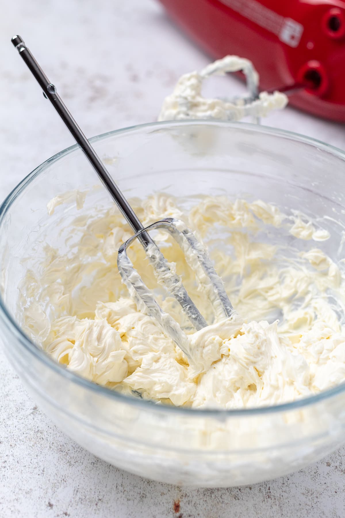 Glass bowl of beaten cream cheese with a mixer beater resting inside and a red hand mixer behind it.
