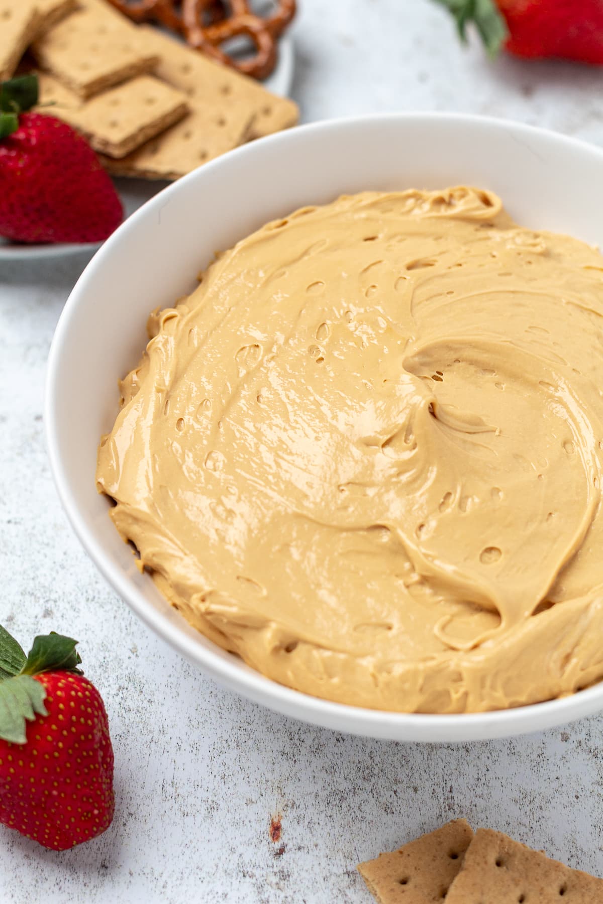 White bowl of creamy light brown dip with strawberries, graham crackers, and pretzels around it.