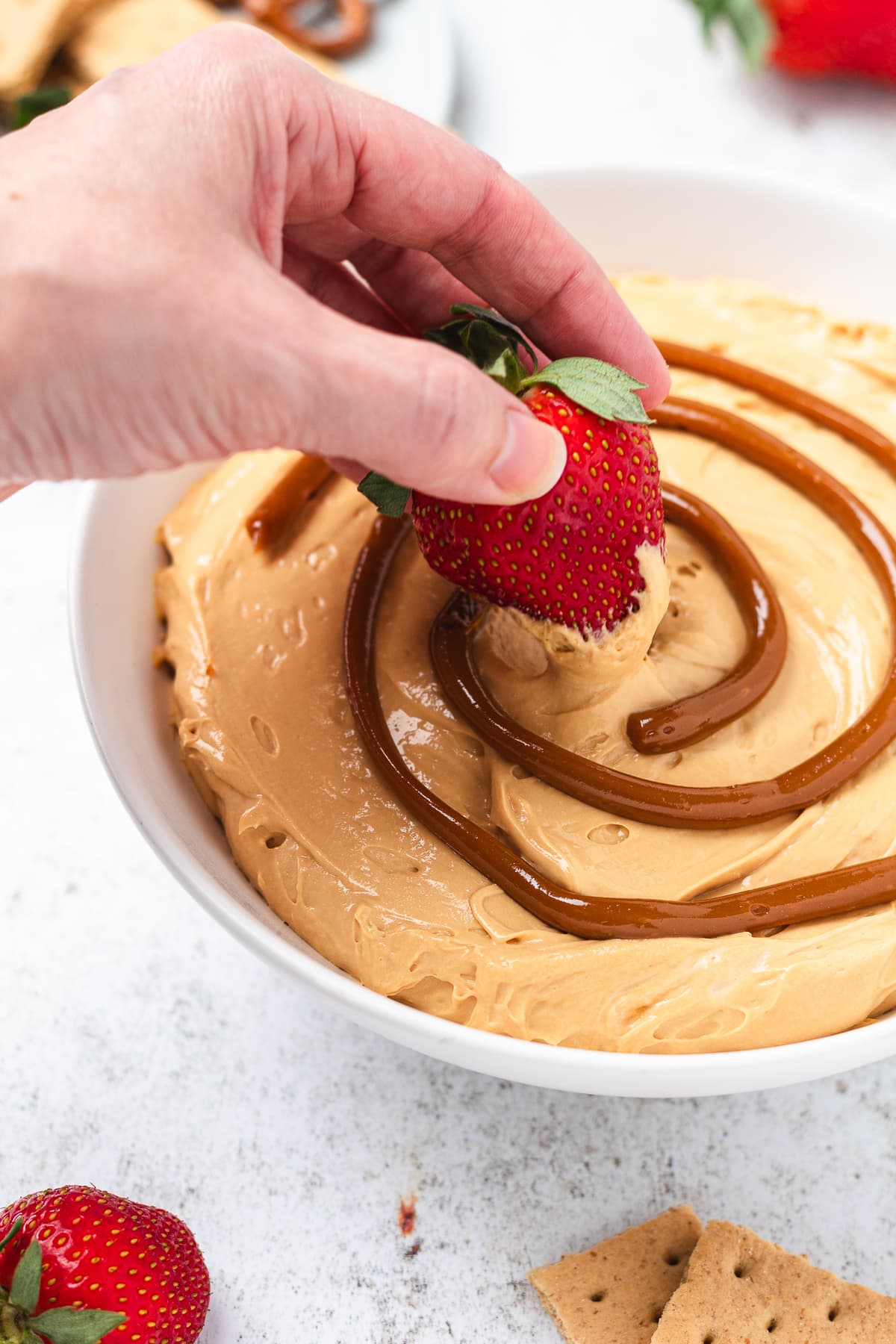 Hand dipping a strawberry into creamy caramel dip with more strawberries, pretzels, and graham crackers nearby.