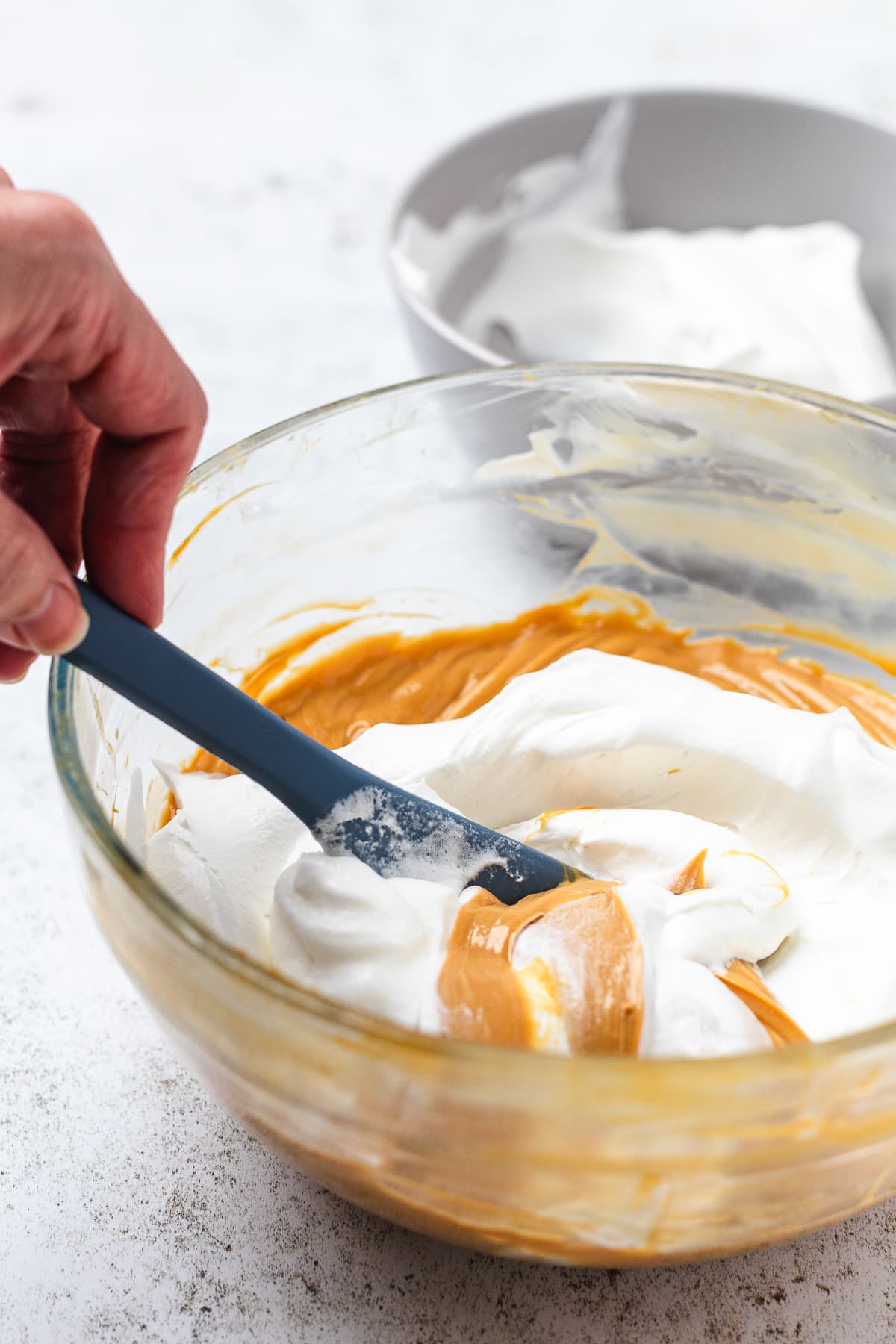 Hand folding a light brown mixture with a blue spatula into a glass bowl of fluffy white mixture.