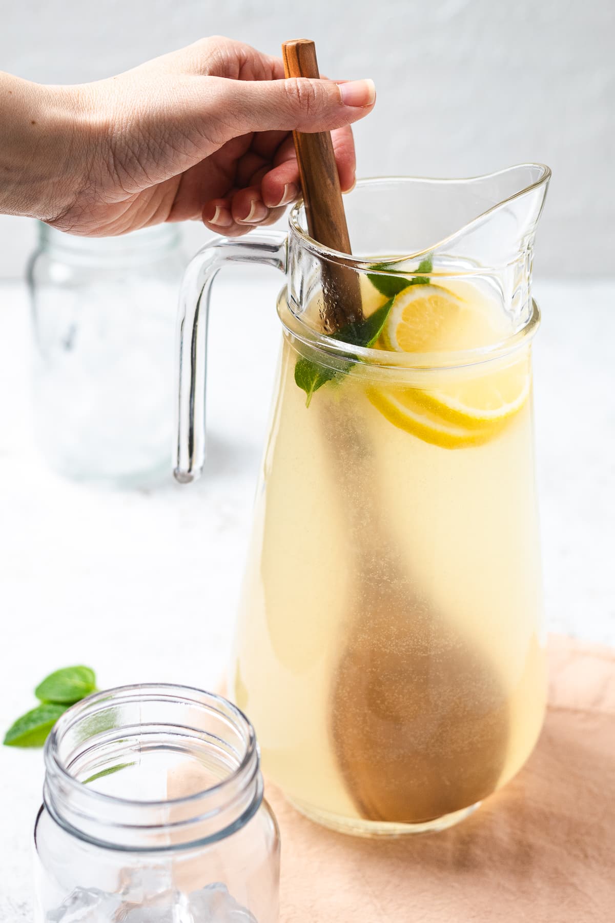 Hand stirring fresh lemonade in glass pitcher with lemon slices and mint leaves.