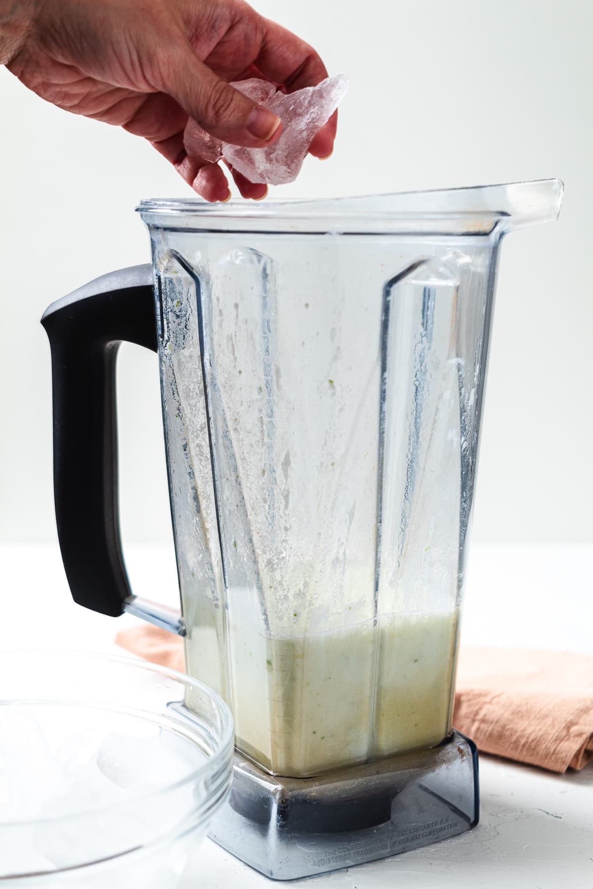 Hand adding ice to blender with creamy lime mixture, pin shows frozen key limeade recipe.