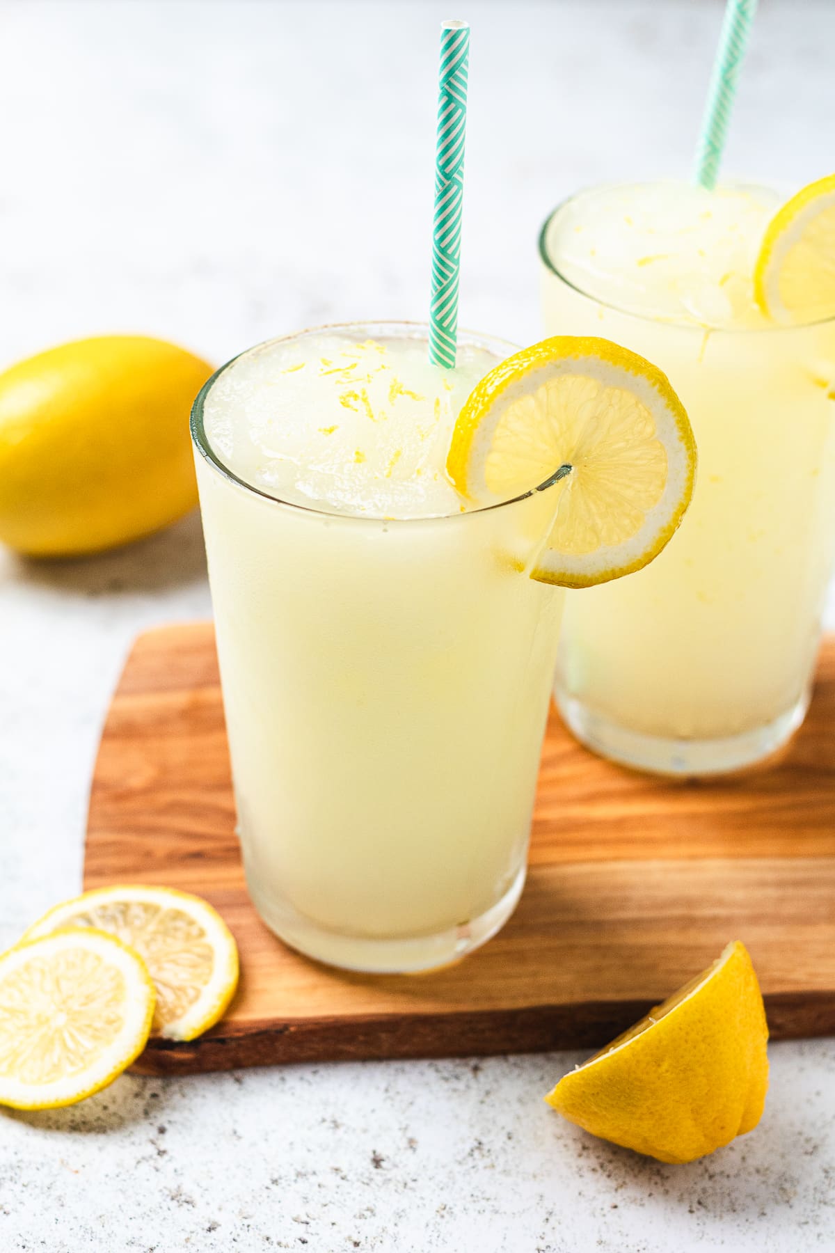Two glasses of frozen lemonade with straws and lemon slices on a wooden board with fresh lemon pieces nearby.