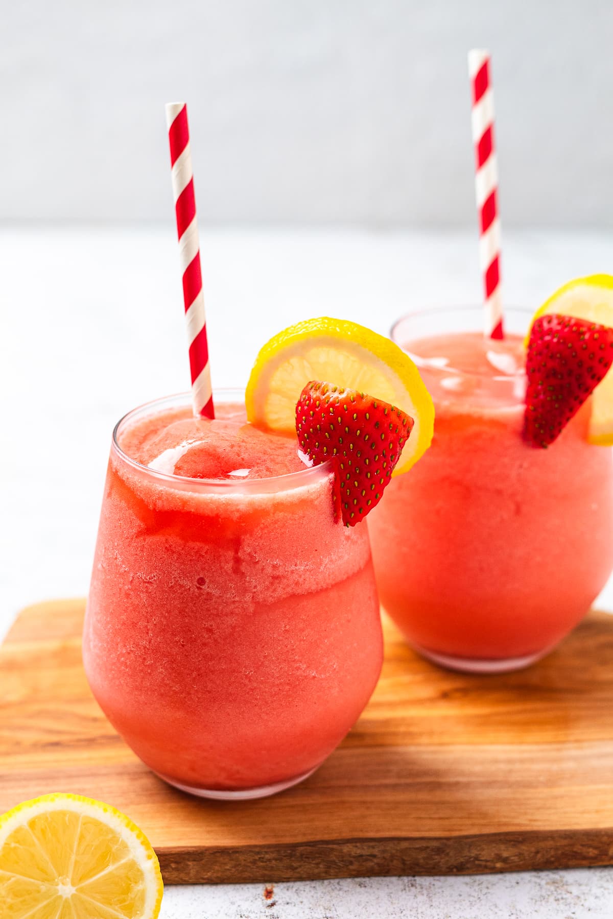 Two glasses of frozen strawberry lemonade with lemon slices and striped straws on wooden board.