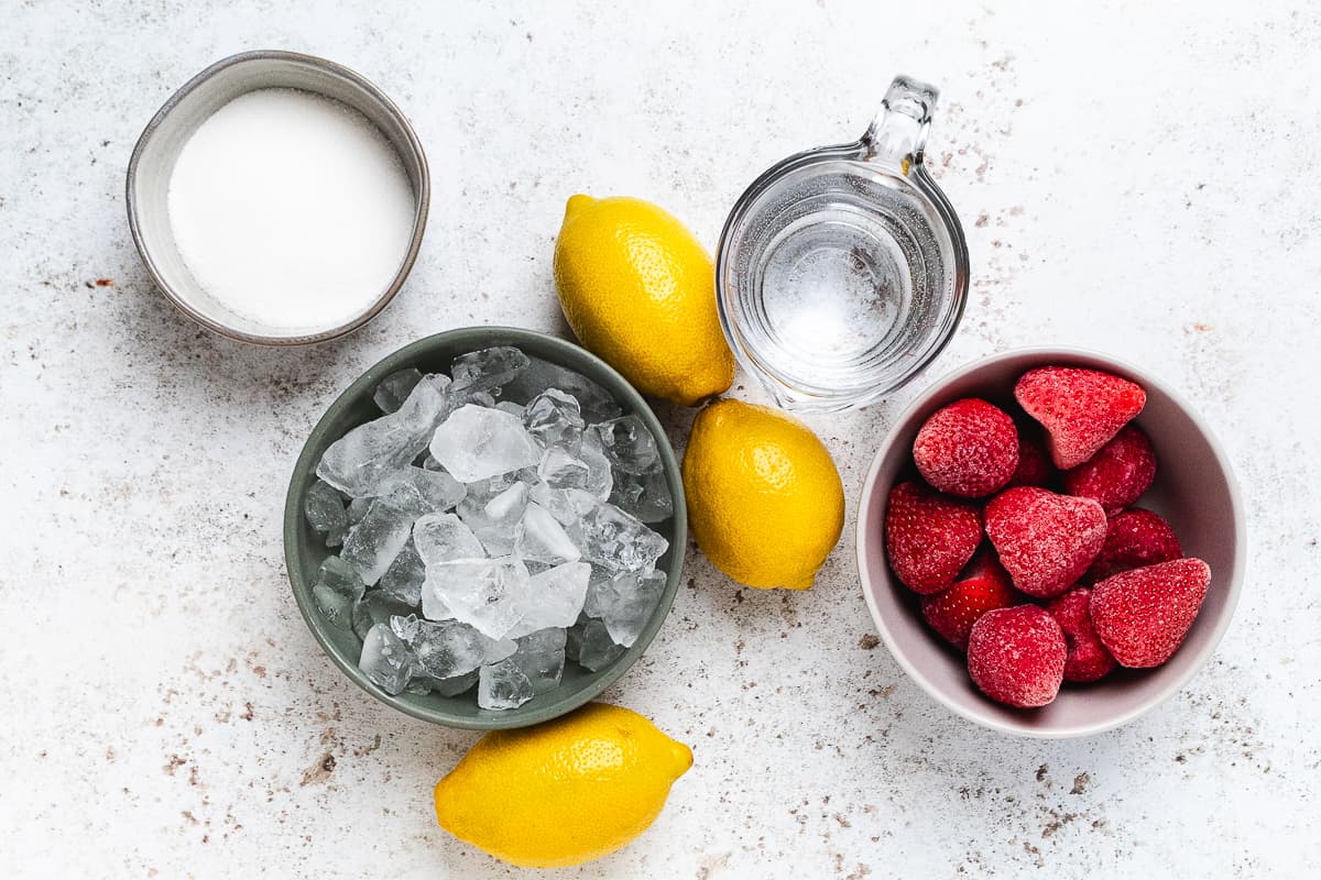 Ingredients for frozen strawberry lemonade: lemons, strawberries, sugar, ice, and water arranged on the surface.