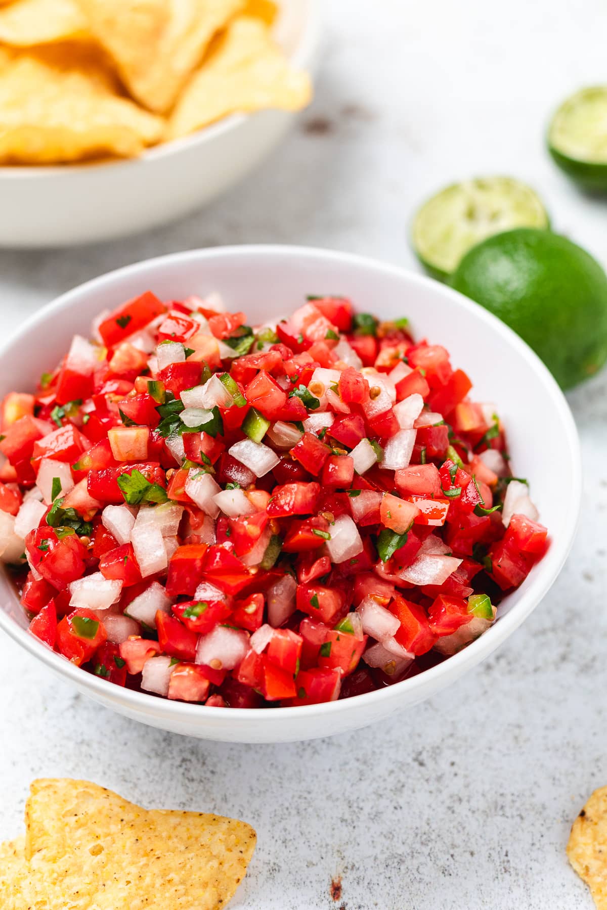 Bowl of pico de gallo with tomatoes, onions, jalapeรฑos, cilantro; chips and halved limes in background.