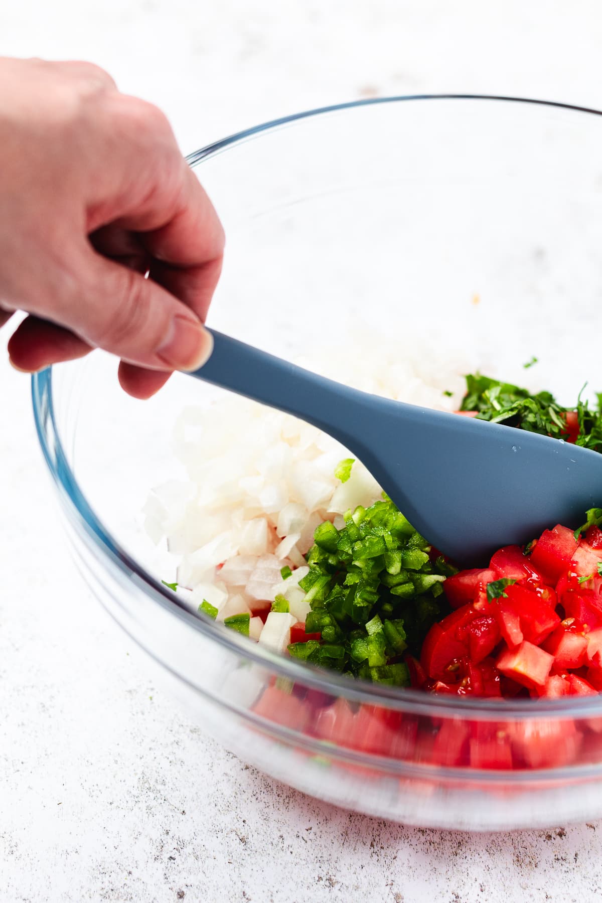 Hand stirring chopped tomatoes, onions, peppers, and cilantro in a glass bowl with a blue spatula.