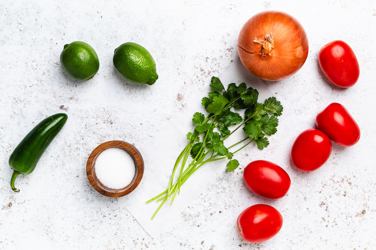 Whole limes, onion, Roma tomatoes, cilantro, jalapeรฑo, and a small bowl of salt on a light surface.