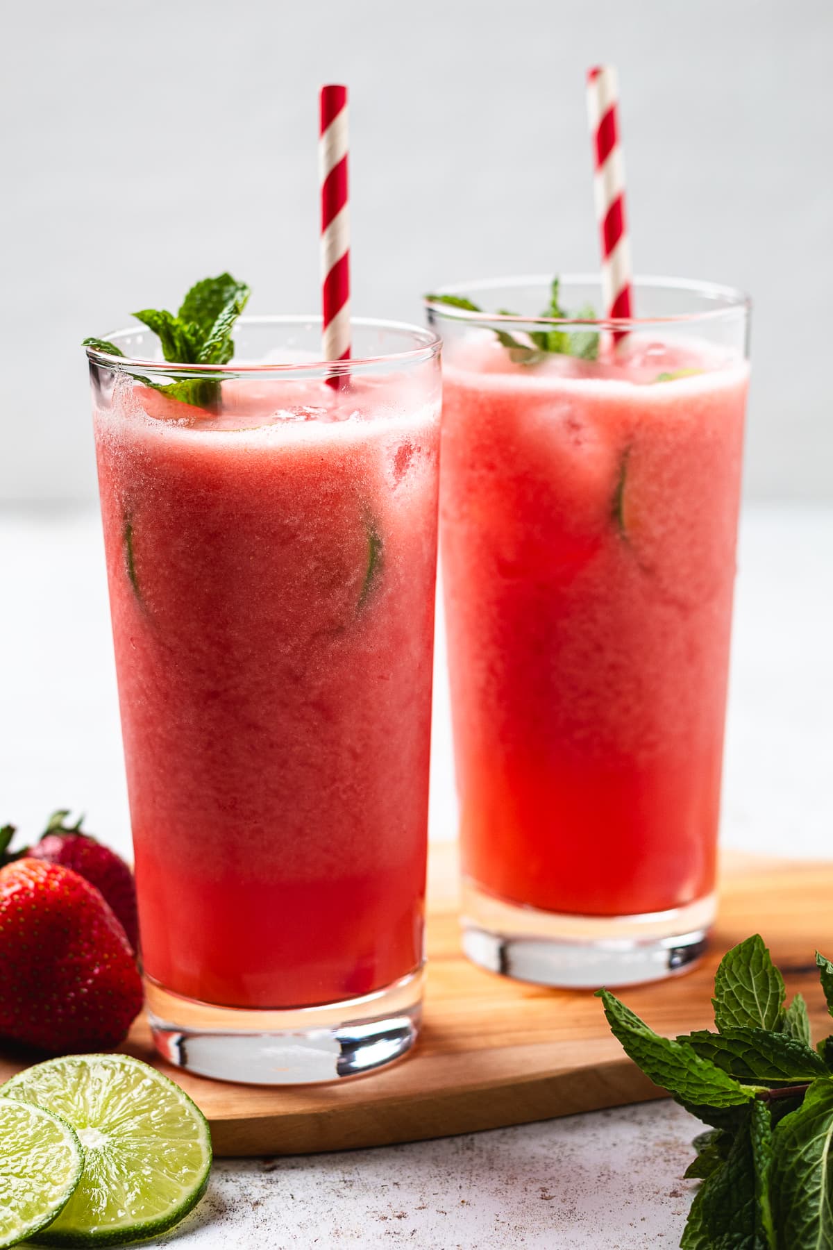 Two glasses of pink red drink with mint and striped straws on a wooden surface with strawberries and limes.