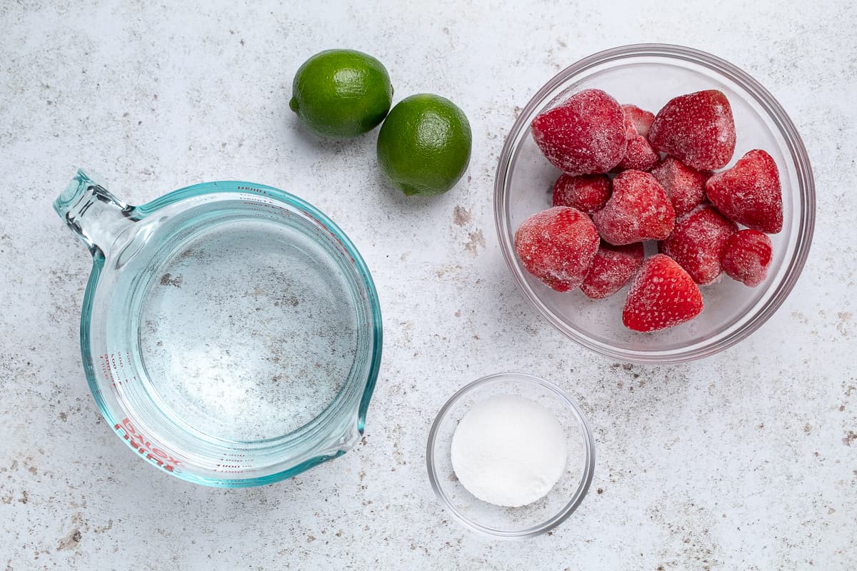 Measuring cup of water with limes, frozen strawberries in a bowl, and sugar in a small dish on a light surface.