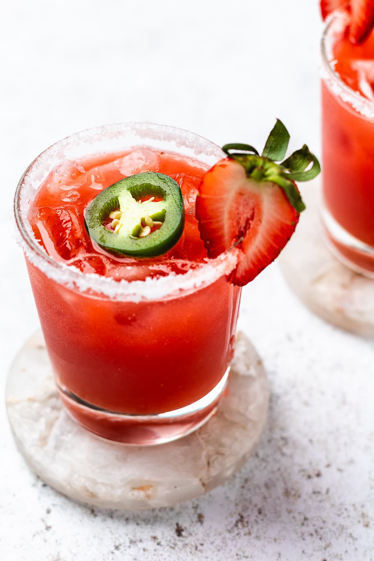 Glass of red drink with ice, salted rim, jalapeño slice, and strawberry with another drink behind it.