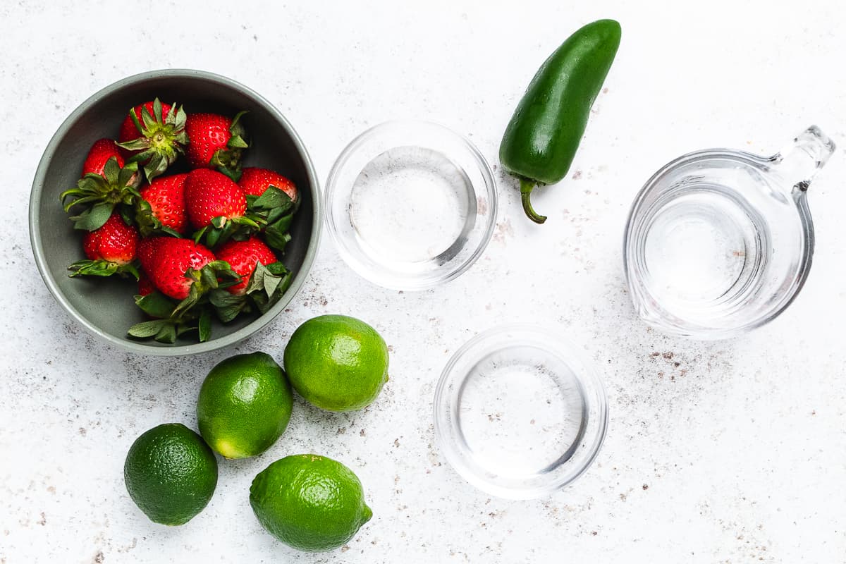 Bowl of strawberries with limes, a jalapeรฑo, and three clear containers of water on a light surface.