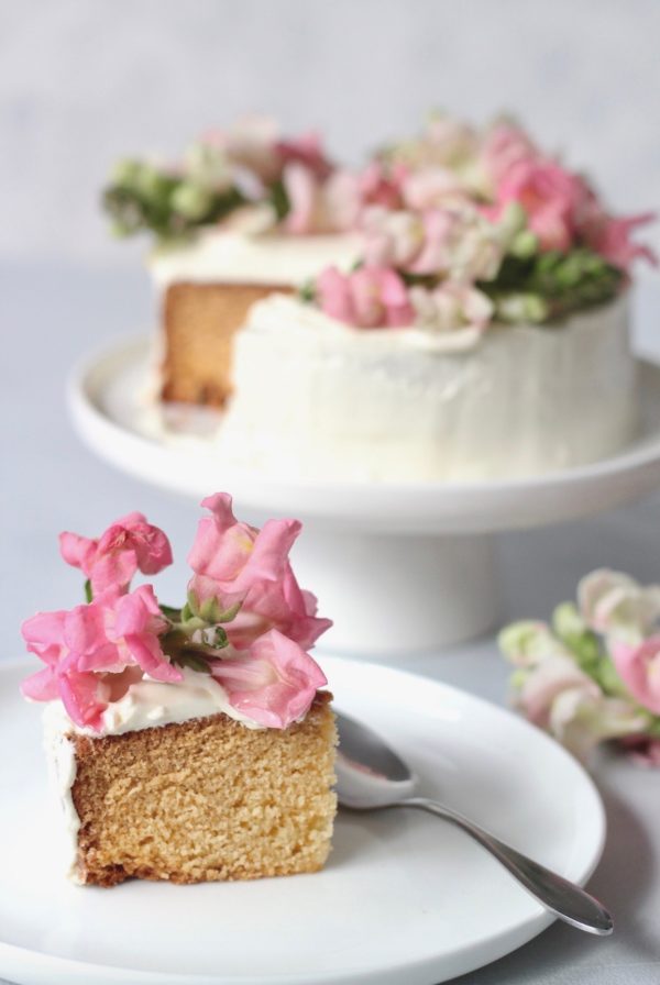 Slice of vanilla cake with white frosting and pink flowers, with whole cake in background.
