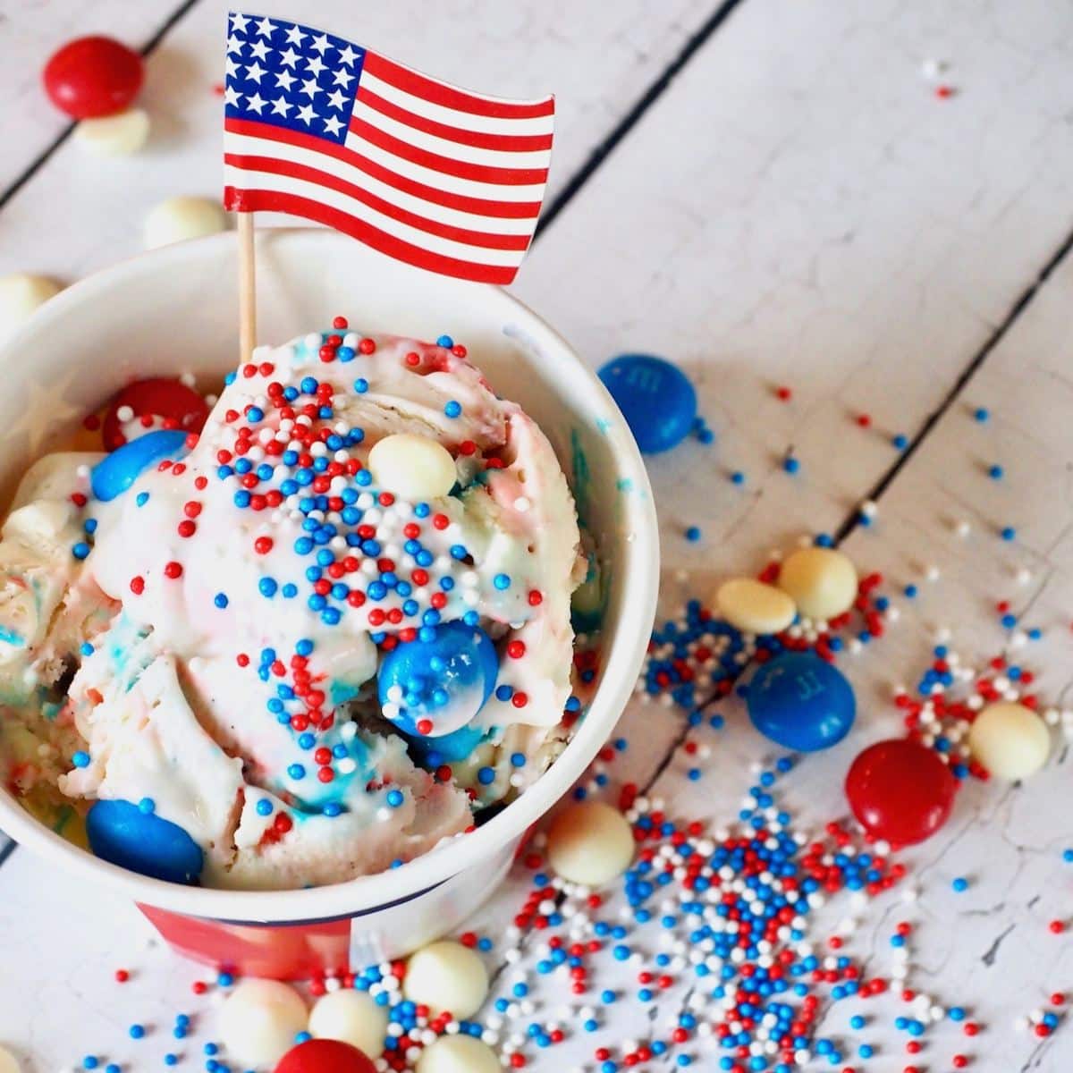 Scoop of vanilla ice cream with red, white, and blue sprinkles and candy, topped with small American flag.