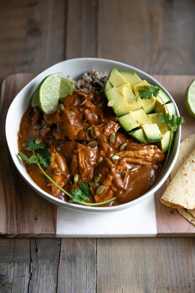 Chicken mole bowl topped with avocado slices, pepitas, and lime wedges.