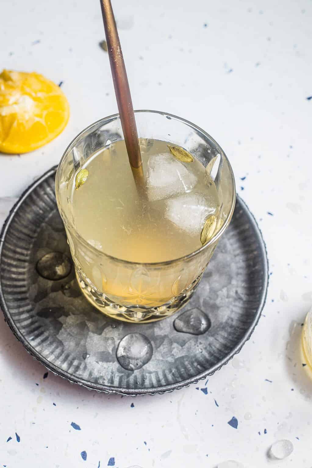 Glass of lemonade with cardamom pods and ice on metal saucer with straw.