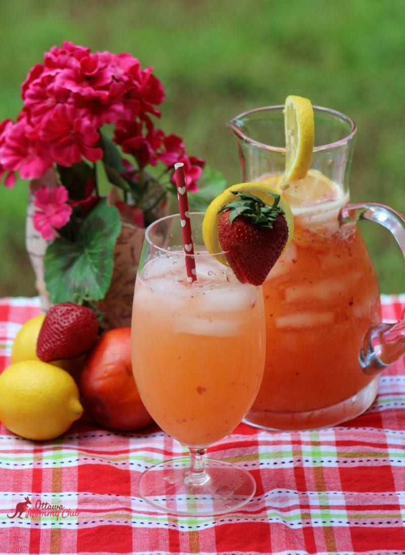Glass and pitcher of strawberry lemonade with lemon slices on red checkered tablecloth.