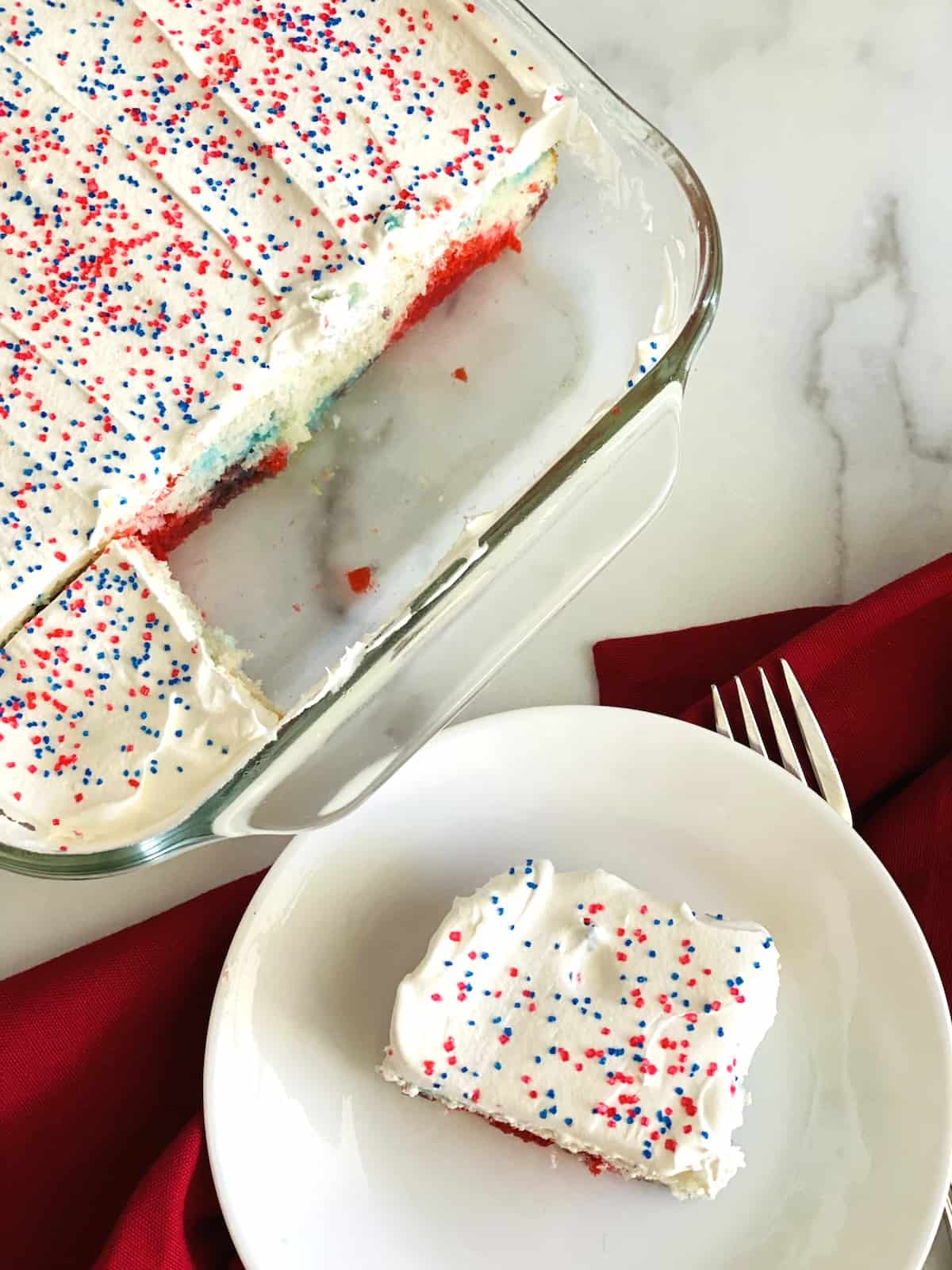 Red, white, and blue poke cake with whipped topping and sprinkles, sliced in glass baking dish.