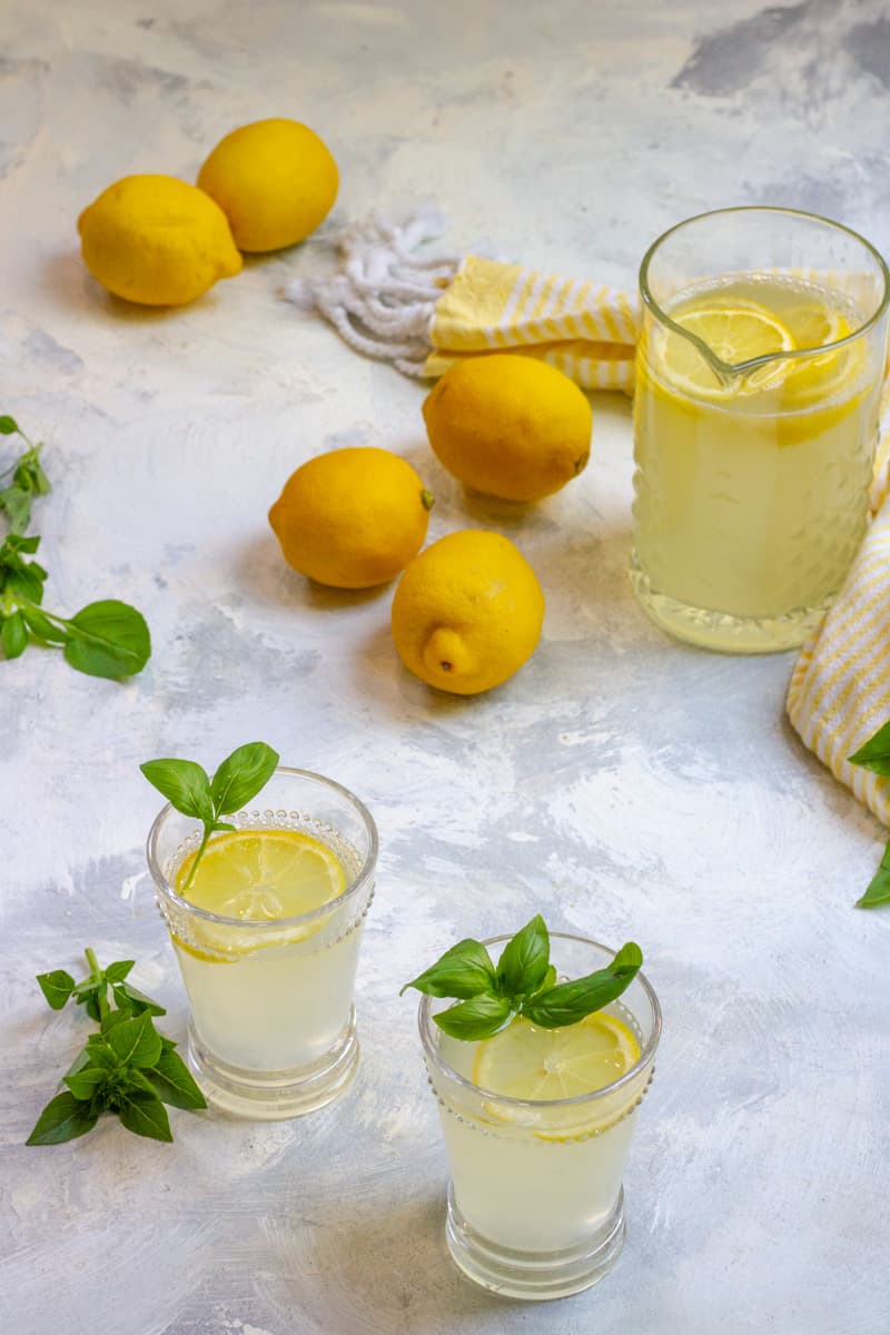 Glasses of lemonade with lemon slices and basil leaves, pitcher and fresh lemons on light surface.