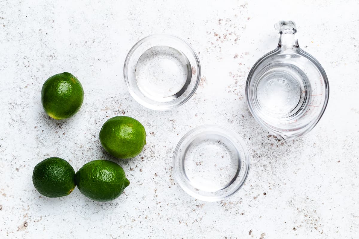 Ingredients for a classic margarita including fresh limes, tequila, and lime juice on a white surface.