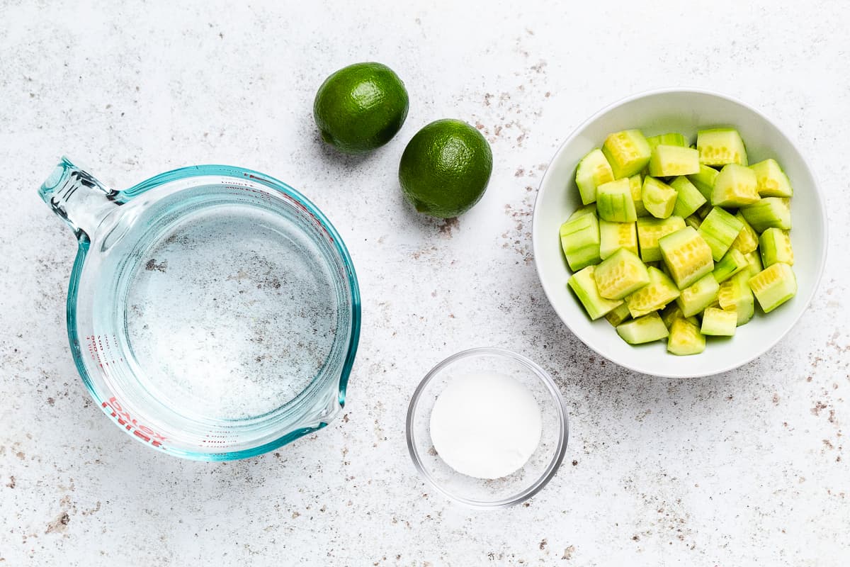 Cucumber agua fresca ingredients including cucumbers, limes, sugar, and water on white surface.