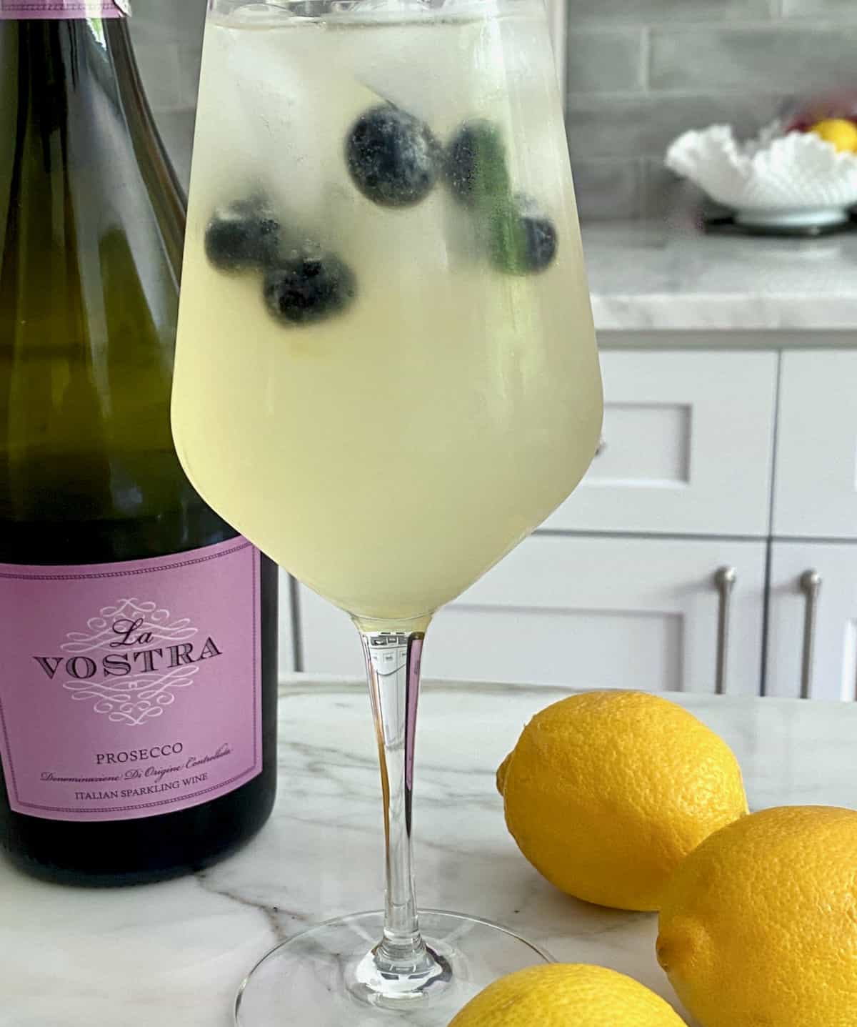 Tall glass of lemonade with blueberries and ice beside bottle of prosecco and whole lemons.