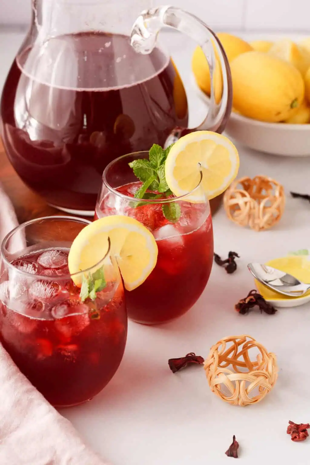 Glasses of hibiscus lemonade with lemon slices and mint, pitcher in background.