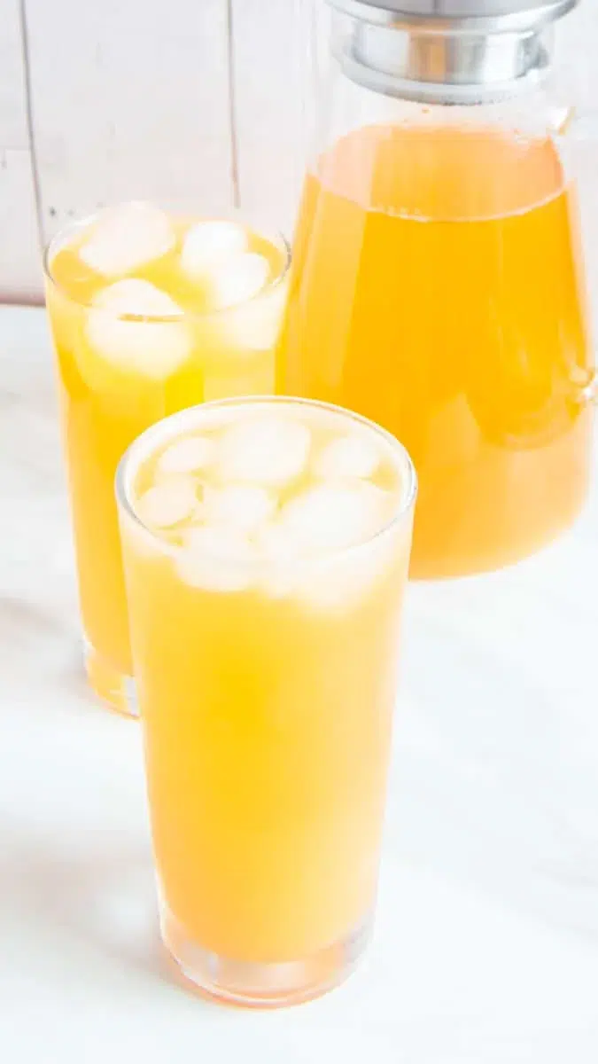 Glasses of iced peach green tea lemonade with ice and pitcher behind.