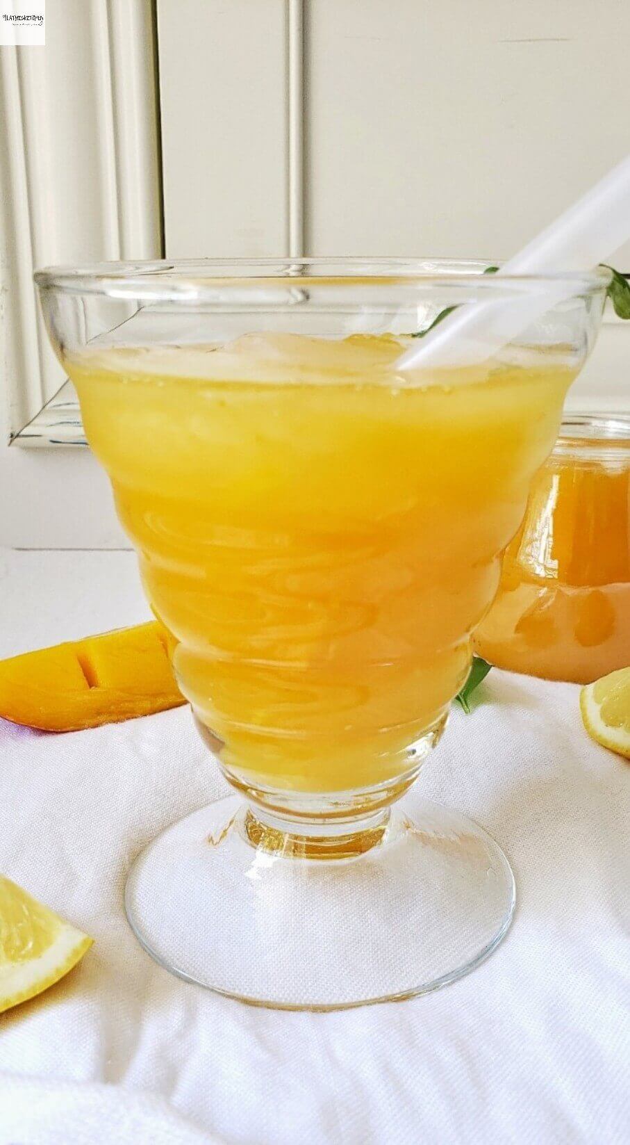 Mango lemonade in large glass with ice, lemon slices, and mango pieces nearby.