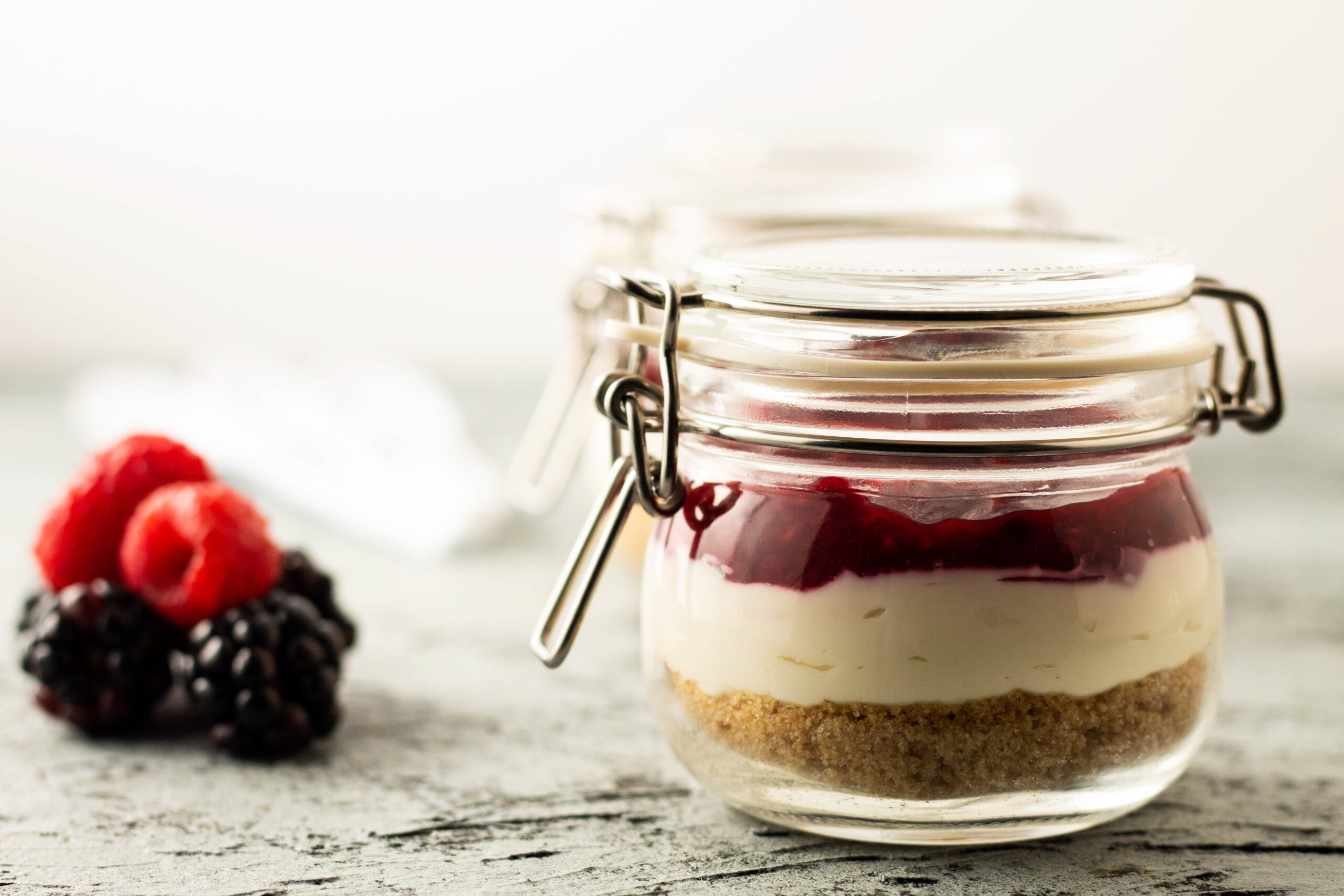 Layered no-bake mixed berry cheesecake in glass jars with berries and spoons on a gray board.
