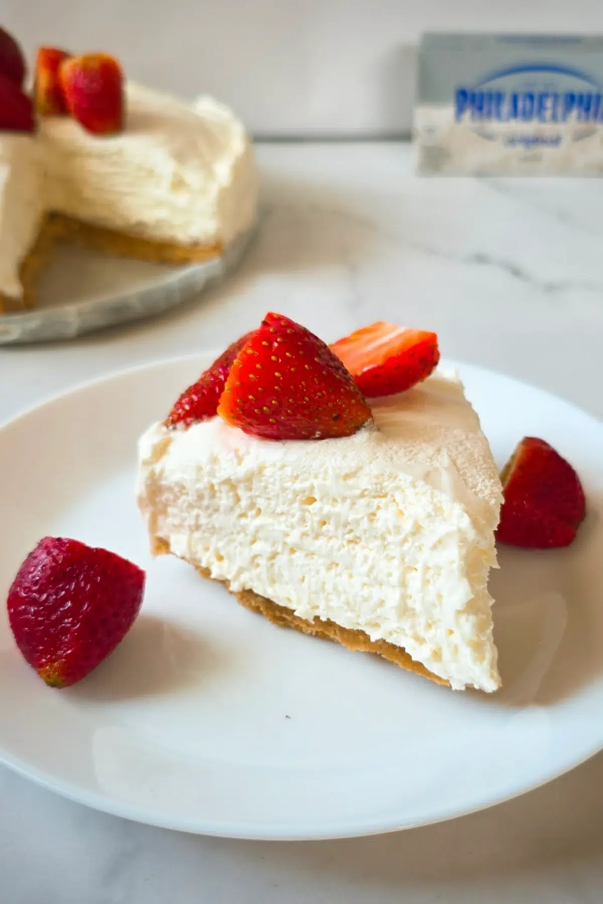 Slice of creamy no bake cheesecake with graham crust topped with fresh strawberries on white plate.