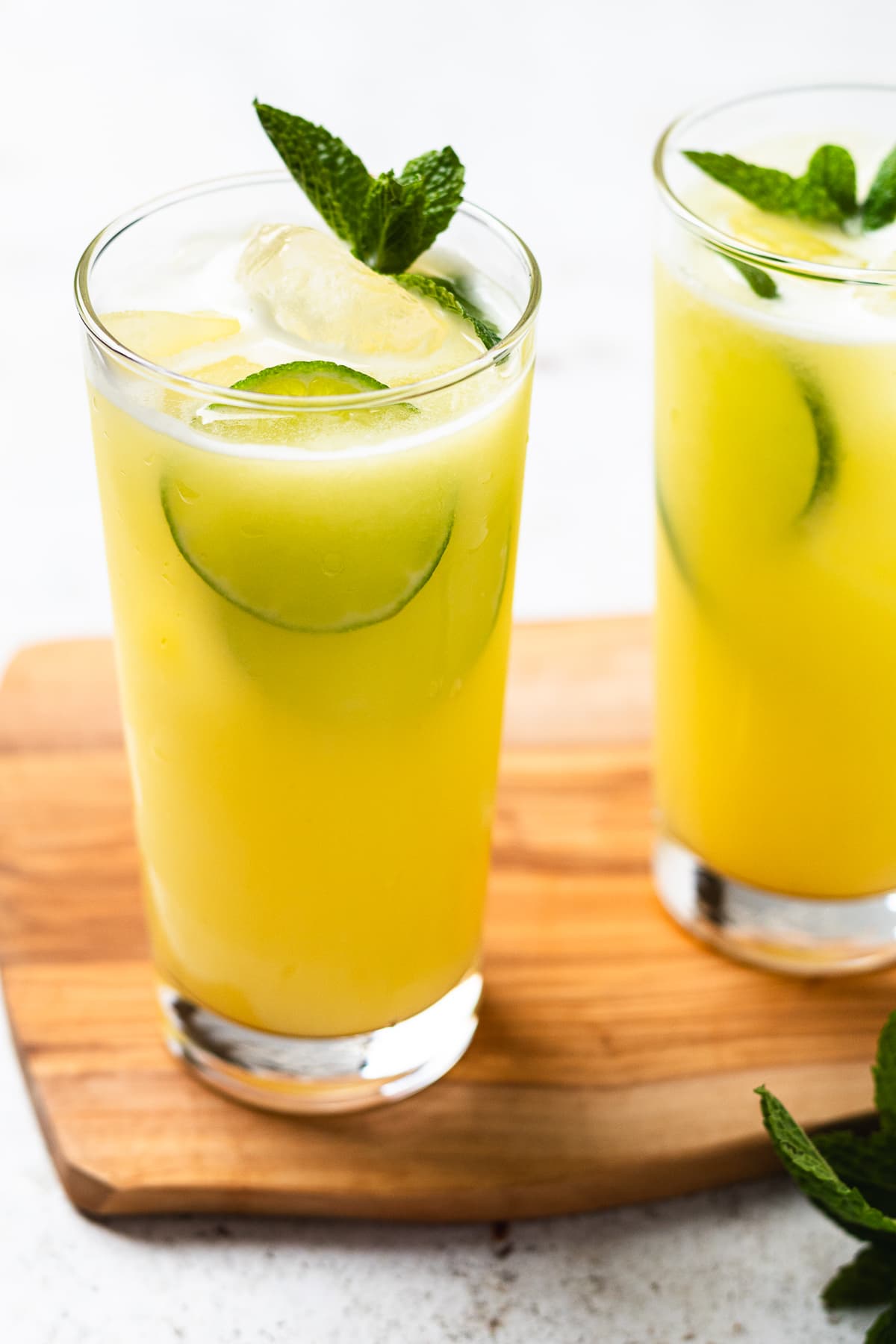 Two glasses of yellow drink with ice, cucumber slices, and mint on a wooden board.