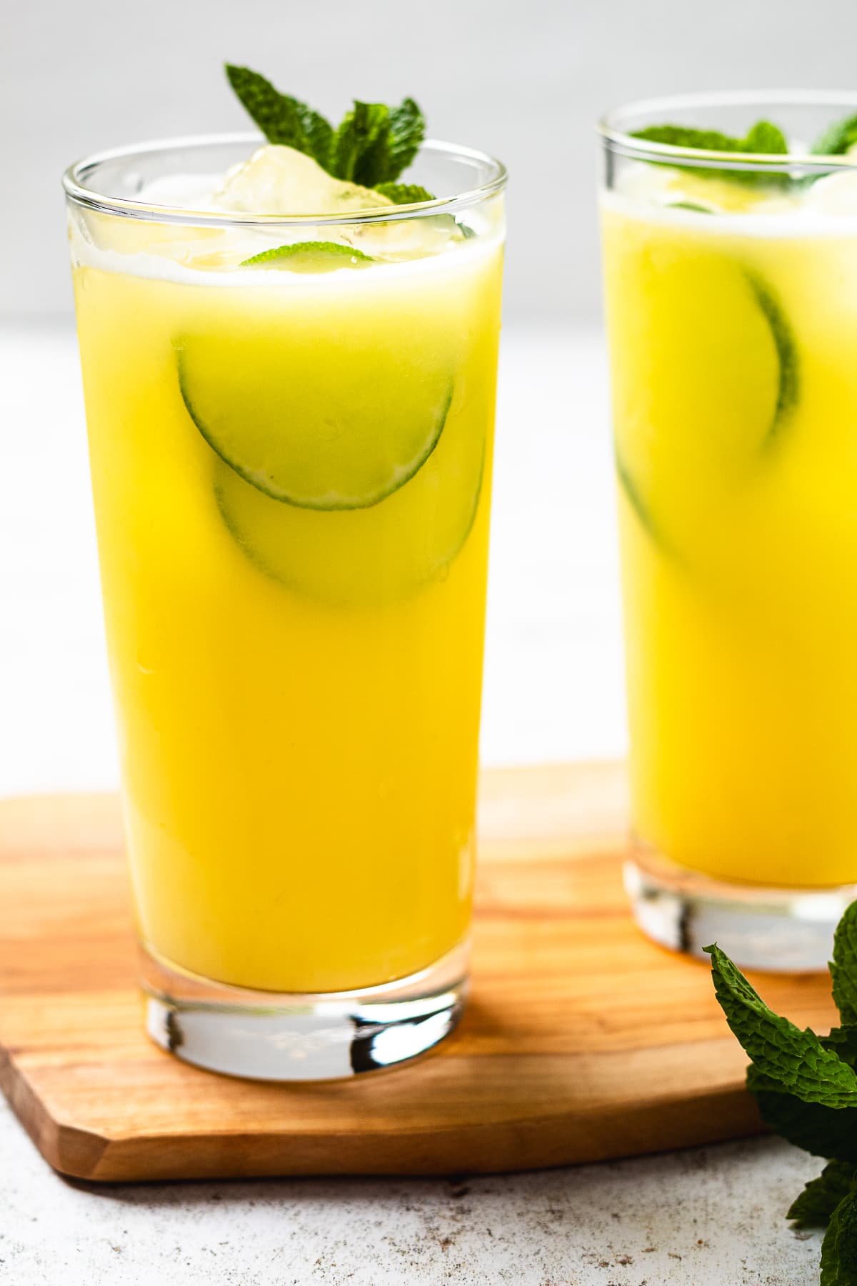 Two glasses of yellow drink with ice, cucumber slices, and mint on a wooden board.