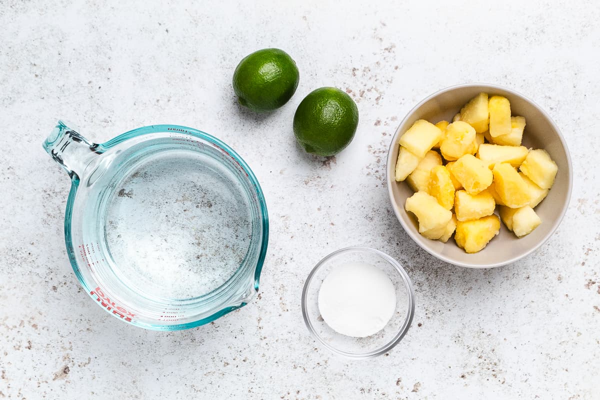 Water in a measuring cup with whole limes, pineapple chunks in a bowl, and sugar in a small dish.
